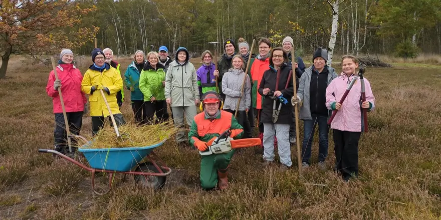 Eine Gruppe von freiwilligen Naturschutzhelferinnen und -helfern steht auf einer Heidefläche und lächelt in die Kamera. Sie halten Werkzeuge wie Rechen, Astscheren und eine Motorsäge; im Vordergrund steht eine Schubkarre voller Schnittgut.